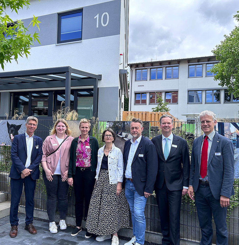 Sieben Personen in Business- und Freizeitkleidung posieren vor einem Gebäude mit Hausnummer 10; Gartenzaun im Vordergrund und blauer Himmel.