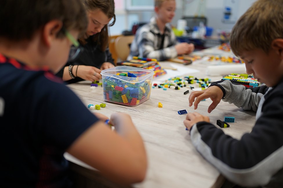 Kinder sitzen an einem Tisch und bauen gemeinsam mit bunten Bausteinen, die in einer transparenten Box auf dem Tisch liegen.