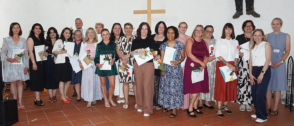 Gruppenfoto: rund 20 Personen in festlicher Kleidung vor einer Kirchenwand mit Kreuz, viele halten Urkunden und einzelne Rosen — feierliche Abschluss‑Szene.
