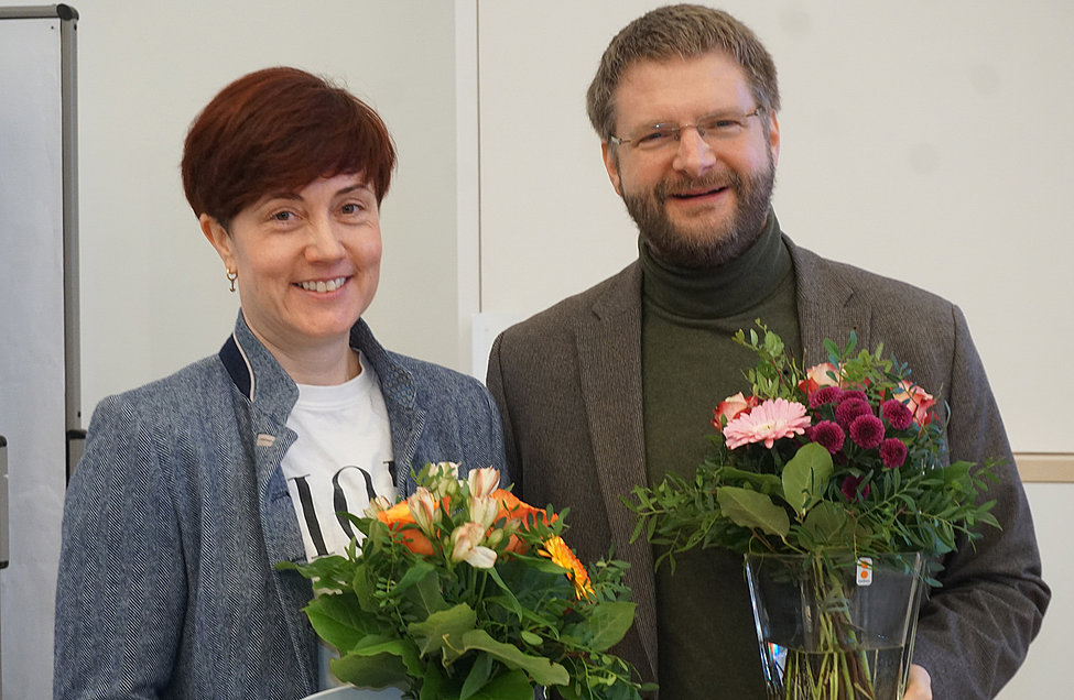 Eine Frau und ein Mann lächeln in die Kamera und halten jeweils einen bunten Blumenstrauß in der Hand. Beide wirken fröhlich und festlich.