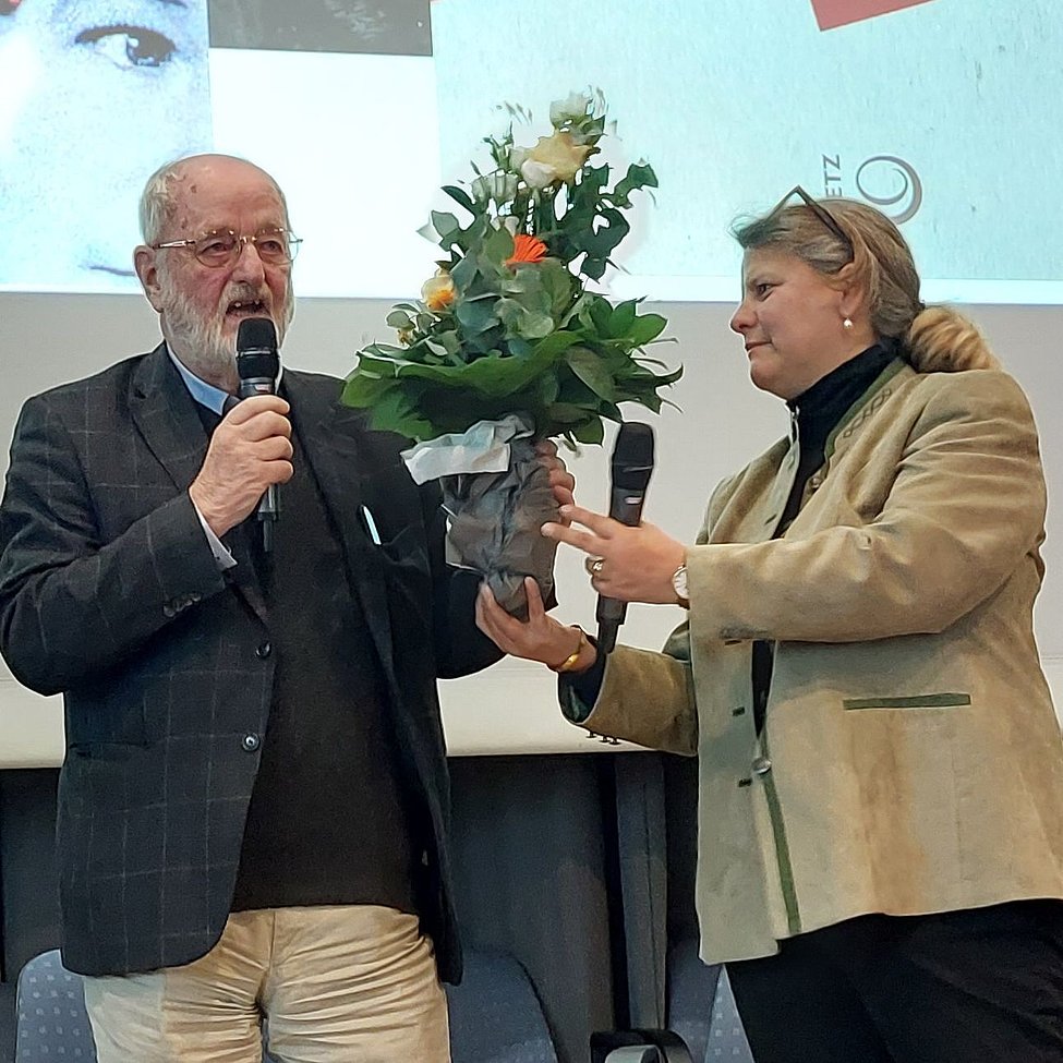 Älterer Mann auf Bühne erhält Blumenstrauß von Frau mit Mikrofon