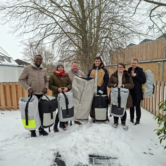 Sechs Personen stehen lächelnd im Schnee und halten Schlafsäcke in den Händen.
