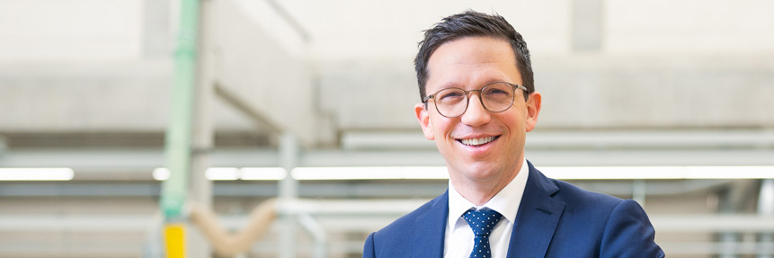 Ein Mann im Anzug steht in einem modernen, hellen Raum und lächelt freundlich in die Kamera. Die Atmosphäre wirkt professionell und offen.